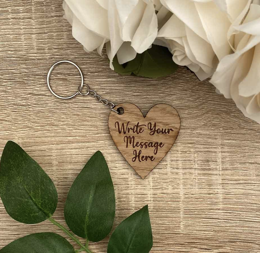 A wooden keyring sits on a table surrounded by flowers. The message on the keyring reads 'Write Your Message Here'