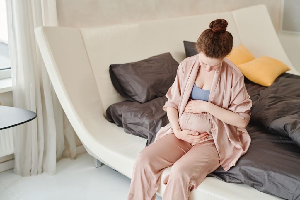 Early Pregnancy Headaches: Causes & Safe Relief 3 A pregnant woman sitting on a bed, embraces her belly in a cozy and modern bedroom setting whilst thinking about early pregnancy symptoms.
