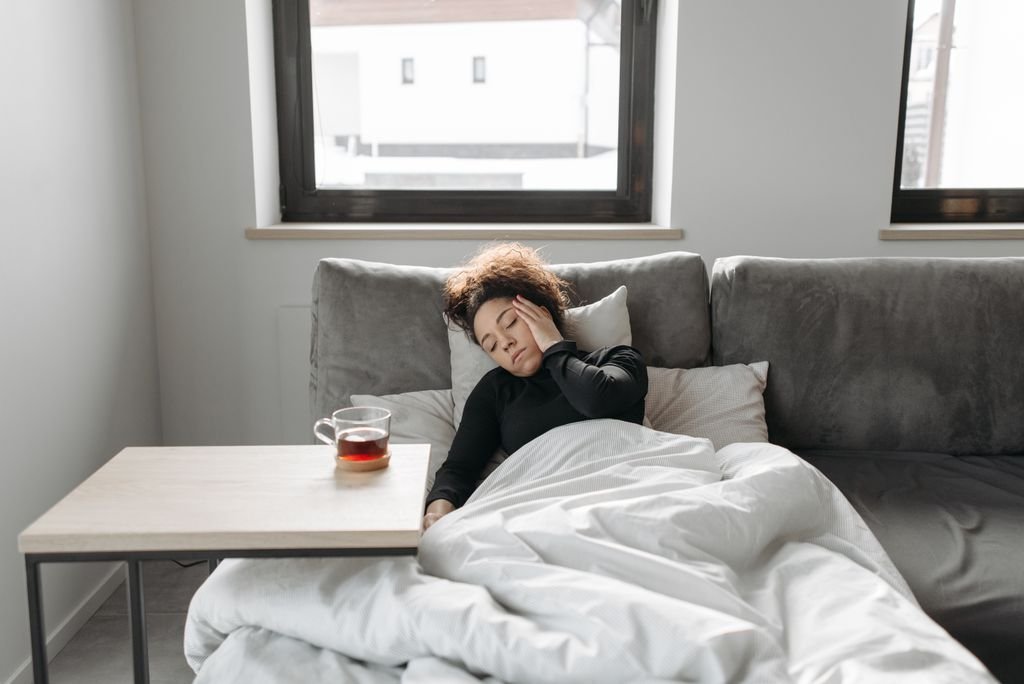 Early Pregnancy Headaches: Causes & Safe Relief 1 A woman rests on a couch indoors, feeling unwell, with an early pregnancy headache and a cup of tea nearby.