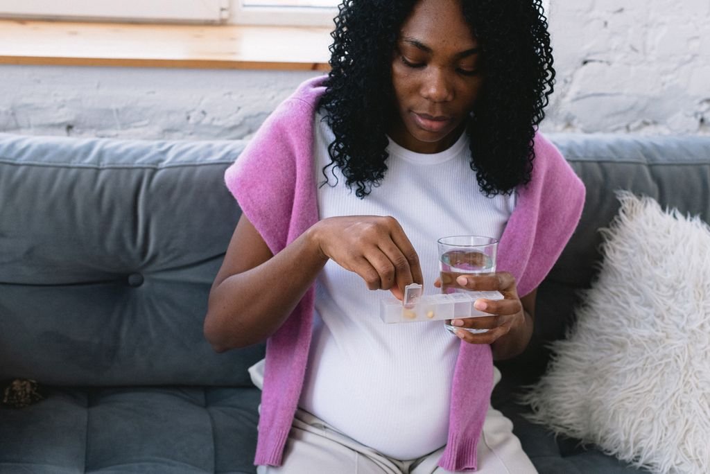 Early Pregnancy Headaches: Causes & Safe Relief 5 Serious pregnant African American female with glass of water taking prenatal vitamins while sitting on comfortable sofa in living room